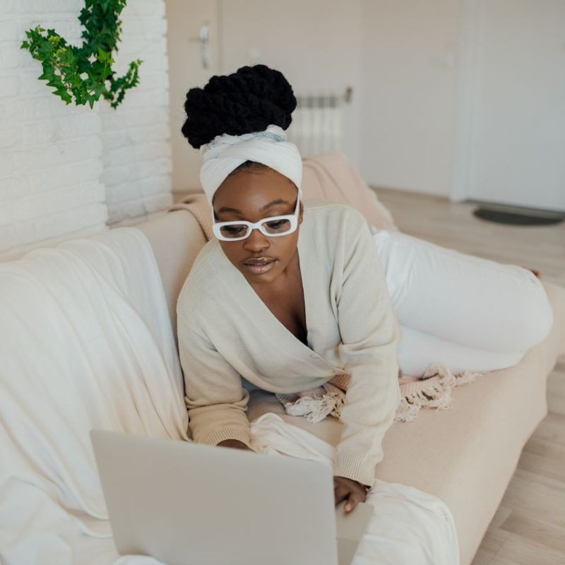 Relaxed African American woman with eyeglasses using laptop on a sofa indoors.